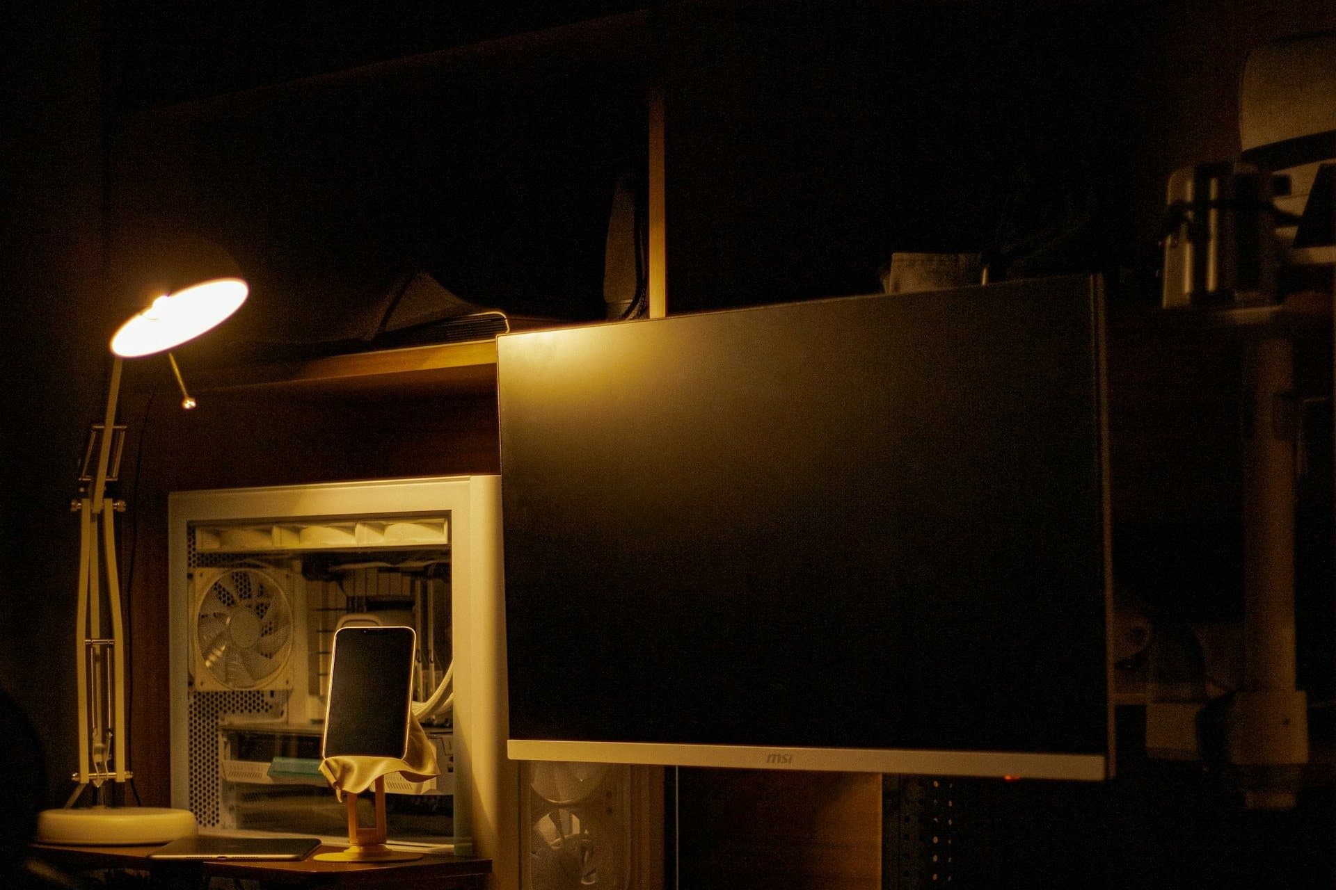 A desk workspace lit by a single warm lamp: a monitor, mechanical keyboard, phone on a stand, and a PC tower
