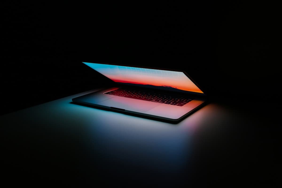 A laptop half open in a dark room, its screen casting a dusk-coloured glow across the desk