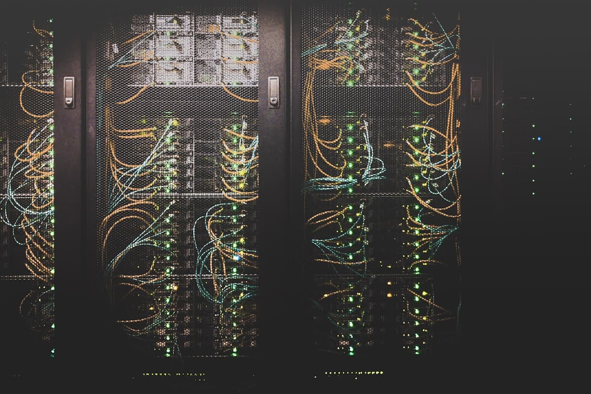 Server racks behind mesh cabinet doors, patch cables lit by rows of green status lights in a dark machine room